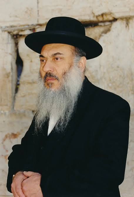 Rabbi Nissim Yagen wearing a black hat and suit beside a stone wall